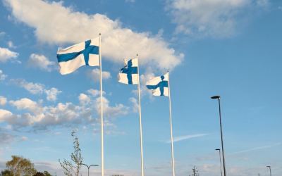 Kolme Suomen lippua liehuu aurinkoisessa ilmassa, sininen taivas pilvineen taustalla.