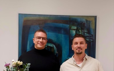 kaksi palkittua miestä seisoo kukkien ja palkinnon kera taulun edessä