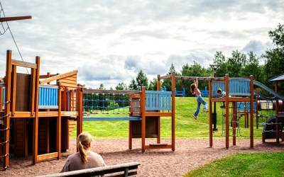 Kuvassa Leijonapuiston leikkiteline ja lapsia leikkimässä. Etualalla nainen istuu penkillä.