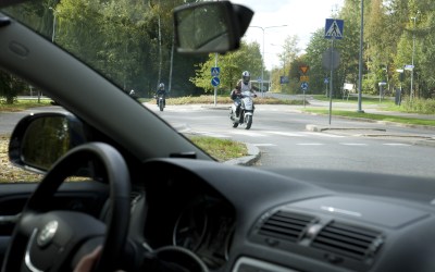Etualalla auton tuulilasi, jonka läpi näkymä liikenneympyrään, jossa ajaa mopoilijoita. Kuvituskuva. Juha Tuomi, Liikenneturva.