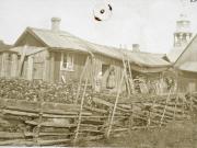 Lindgrenin mökki, pieni puutalo Baltzaborgissa. Riukuaita rajaa kasvimaata, ja pihalla seisoo kaksi naista. Taustalla kohoaa vanhan kirkon torni – tuttu maamerkki kaupunkimaisemassa. Kuva Uudenkaupungin museo. Baltzarborg. Pieni huonokuntoisen näköinen puinen asuinrakennus, ns. Lindgrenin mökki. Etualalla riukuaita, kasvimaa, kaksi naista rakennuksen edustalla, taustalla vanhan kirkon torni.