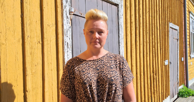 Paula Kesäläinen seisoo keltaisen puutalon edessä ja katsoo päättäväisesti kohti.