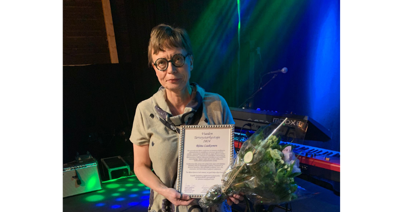 Vuoden terveystarkastajaksi 2024 valittu Riitta Laaksonen pitelee kunniakirjaa ja kukkakimppua.