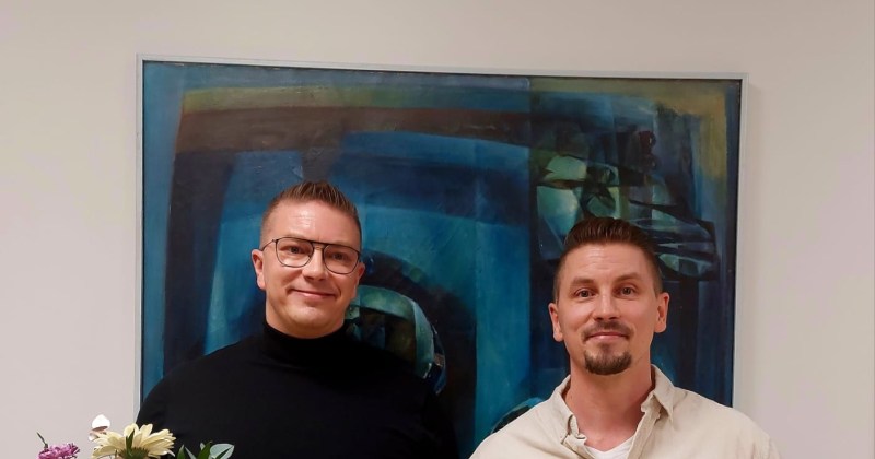 kaksi palkittua miestä seisoo kukkien ja palkinnon kera taulun edessä