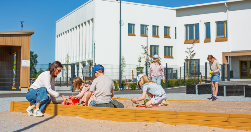 Aikuinen ja lapsia leikkimässä hiukkalaatikolla aurinkoisena kesäpäivänä.