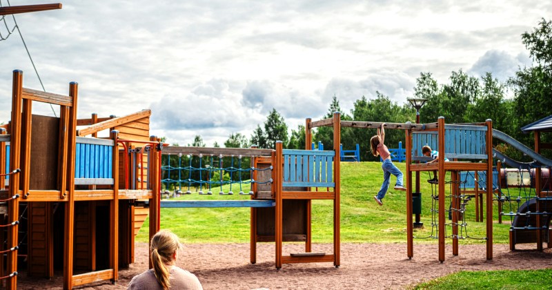 Kuvassa Leijonapuiston leikkiteline ja lapsia leikkimässä. Etualalla nainen istuu penkillä.