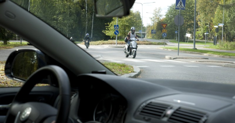 Etualalla auton tuulilasi, jonka läpi näkymä liikenneympyrään, jossa ajaa mopoilijoita. Kuvituskuva. Juha Tuomi, Liikenneturva.