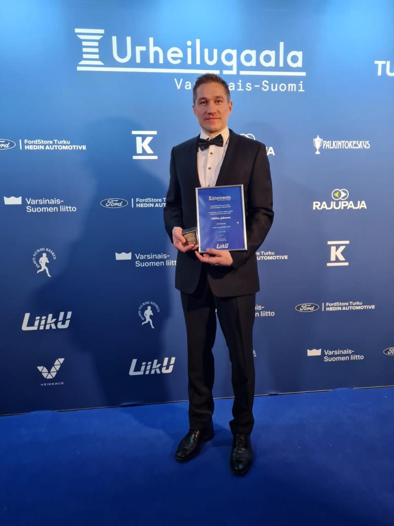 mies puvussa pitelee palkintoa ja kunniakirjaa urheilugaalan taustatekstillä